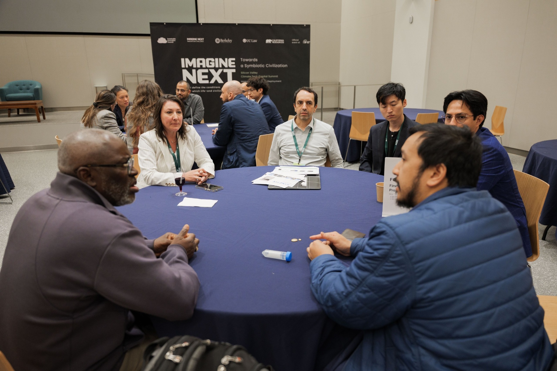 Capital Day Curated Capital Matchmaking Sessions wide shot at UC Law SF — founders and investors in conversation around blue round tables beneath the Imagine Next 'Towards a Symbiotic Civilization' banner