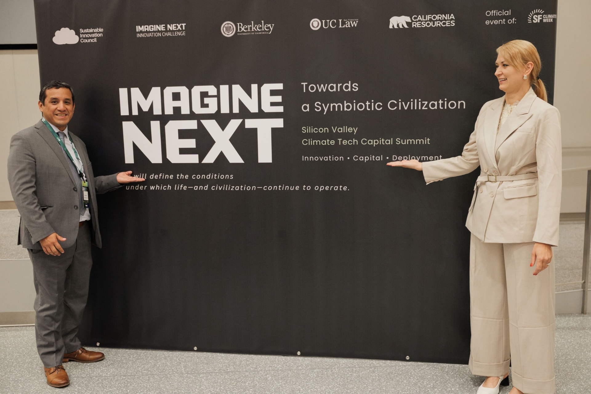 Imagine Next 'Towards a Symbiotic Civilization' Capital Summit backdrop at UC Law SF — side view of two team members presenting the official SF Climate Week sponsor wall