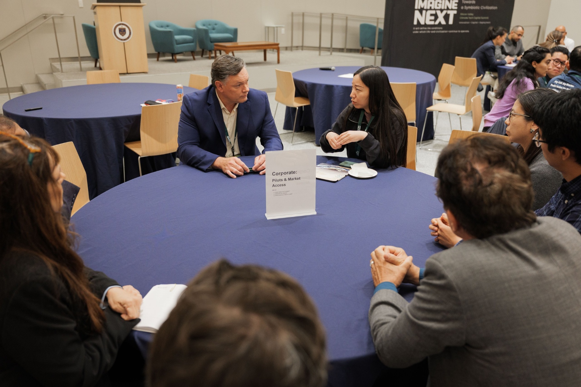 Capital Day Curated Capital Matchmaking Sessions wide shot of the 'Corporate: Pilots & Market Access' table with delegates in conversation beneath the Imagine Next banner at UC Law SF