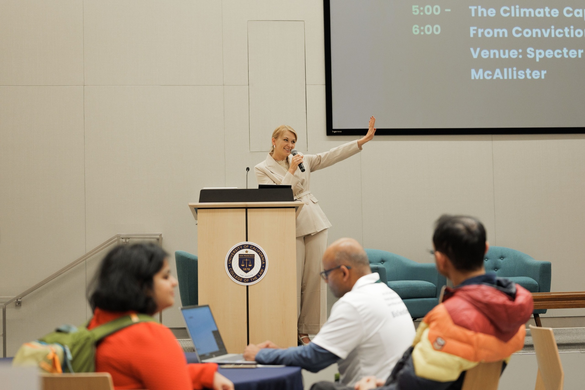 Capital Day speaker at the UC Law SF podium gesturing toward the screen during the 'Climate Capital — From Conviction' session at the Specter McAllister venue