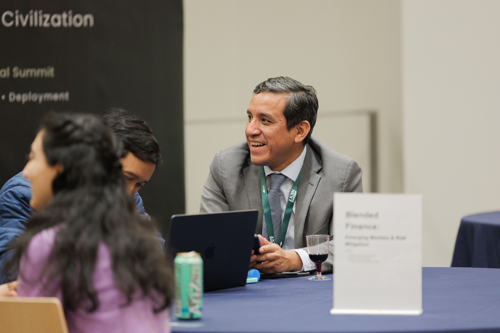 Capital Day Curated Capital Matchmaking Session — delegate in a grey suit smiling in conversation at the 'Blended Finance: Emerging Markets & Risk Mitigation' table beneath the Imagine Next backdrop at UC Law SF